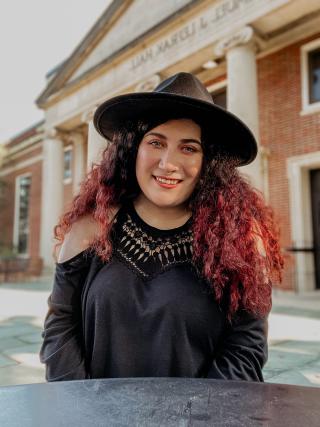 A photo of Xandie Wood smiling at the camera. She is wearing a black wide brimmed hat and a decorative black shirt. She is sitting in front of LeFrak Hall on campus.